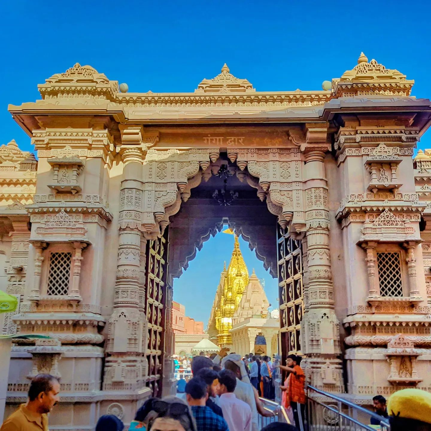 A carved sandstone gateway with the gold spire of the temple seen through its arch, and devotees walking in